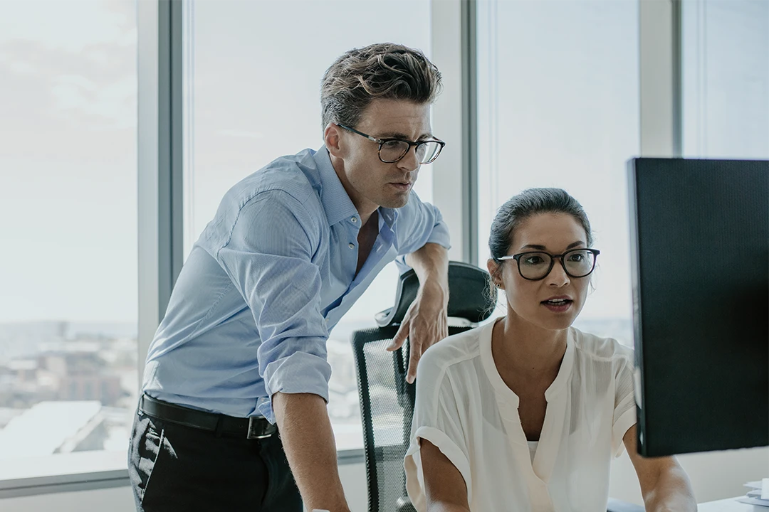 Two colleagues wearing glasses work together at a computer in a modern office with large windows and city views.