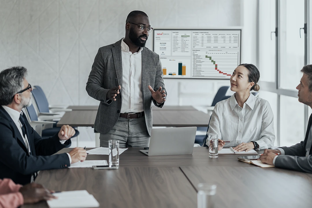 Four people in business attire sit around a conference table. One man stands and speaks, while the others listen. A laptop and papers are on the table, and a presentation with charts is displayed on a screen in the background.