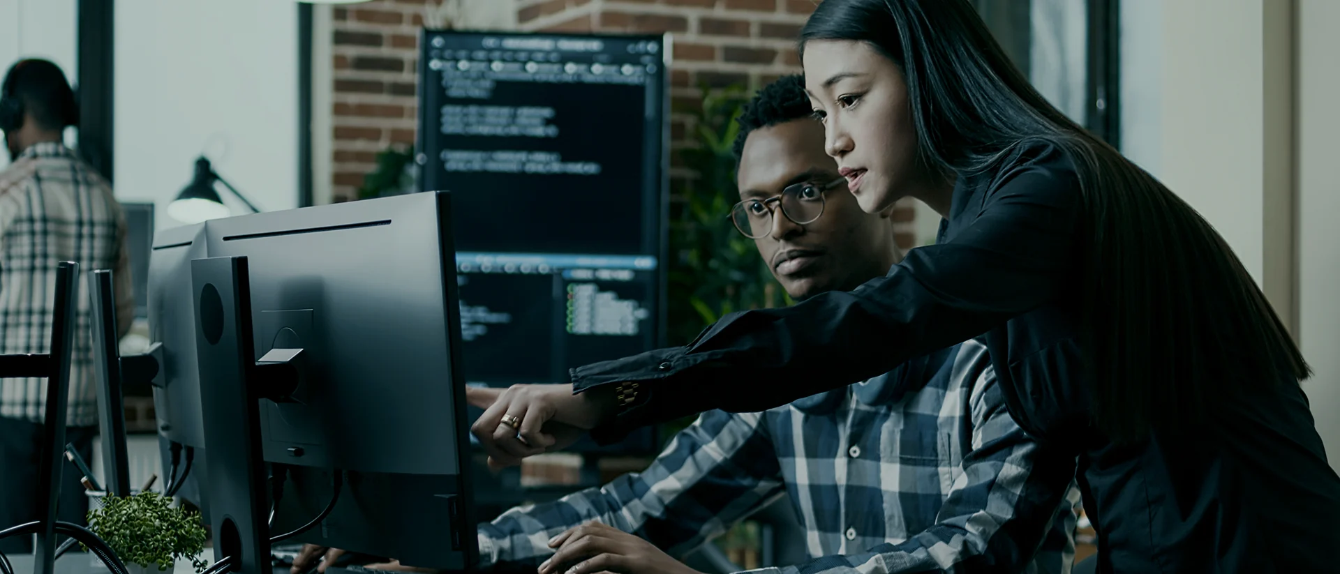 Two people working at a computer, one sitting and typing while the other stands and points at the monitor. Computer code is displayed on screens in the background.