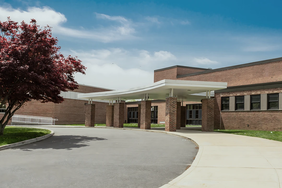 A modern school entrance with brick walls, large windows, and a covered walkway; a tree with red leaves stands to the left under a blue sky with some clouds.