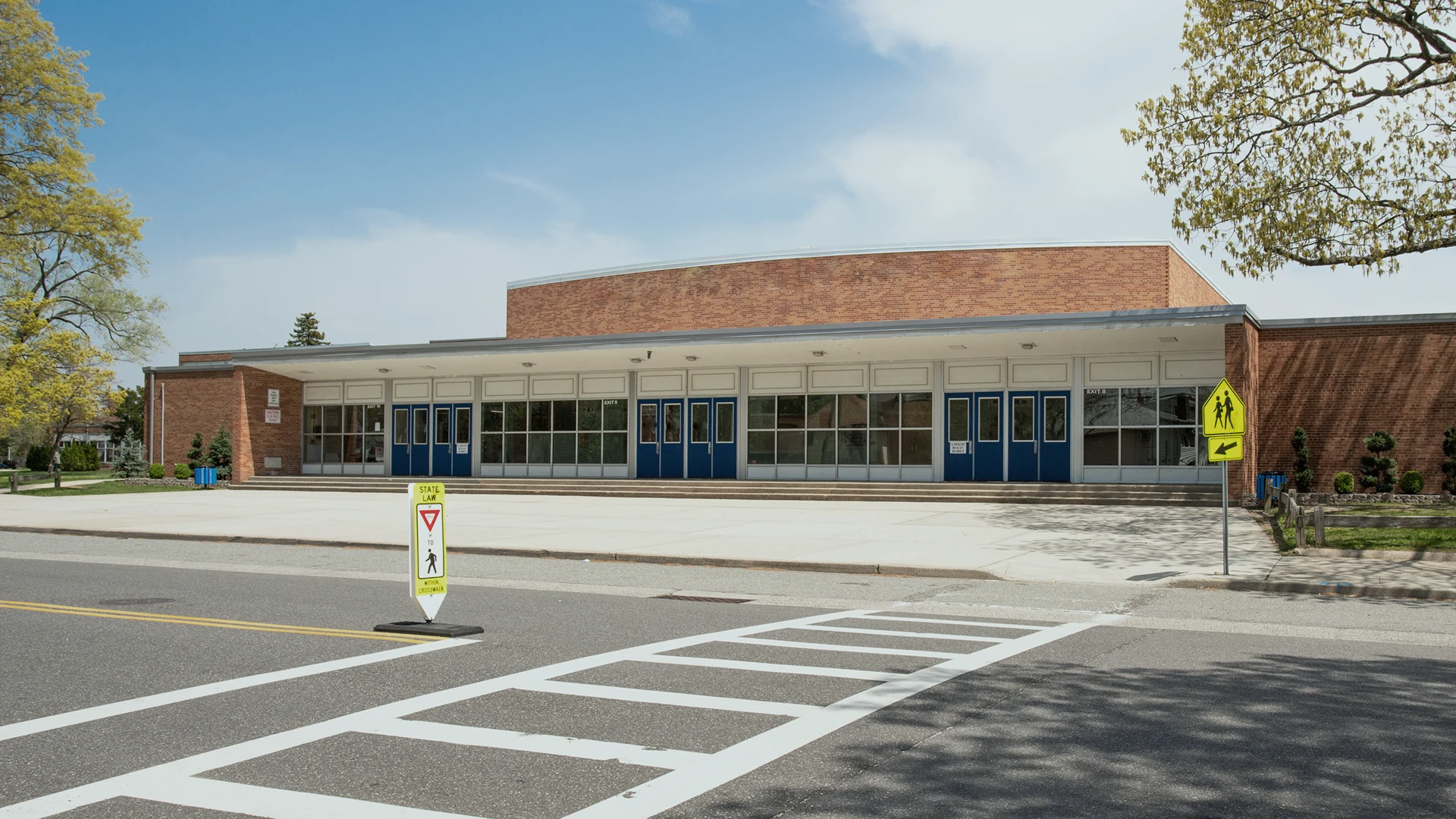 A brick and glass school building with blue doors, large windows, and a flat roof. A crosswalk and a yield sign are visible in front, under a clear sky.