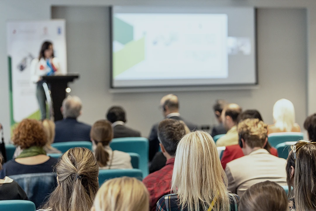 A group of people sits facing a speaker at a podium in a conference room, watching a presentation projected on a screen at the front.