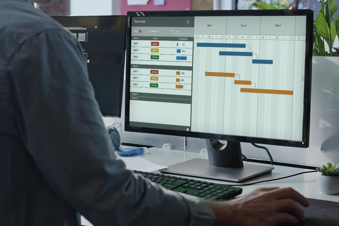 A person sits at a desk using a desktop computer, viewing a Gantt chart and task management software on dual monitors, likely organizing project tasks and timelines.