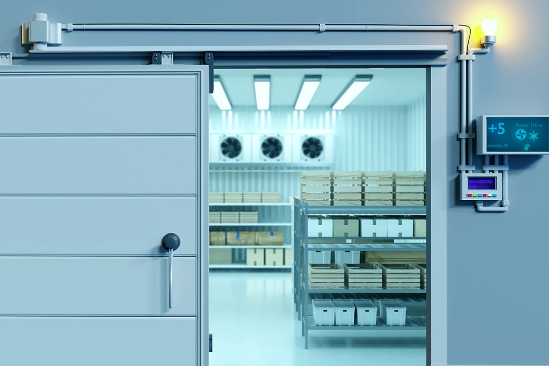 A cold storage room with open door, shelves holding boxes, cooling fans on the back wall, a digital temperature display showing +5°C, and a yellow warning light above the entrance.