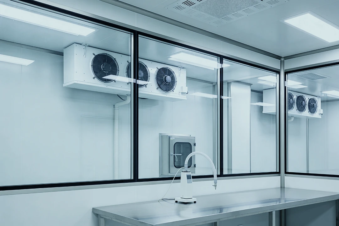 A clean, modern laboratory room with glass windows, stainless steel work surfaces, air filtration units on the walls, and ventilation equipment. The room appears sterile and well-lit, with no people or clutter visible.