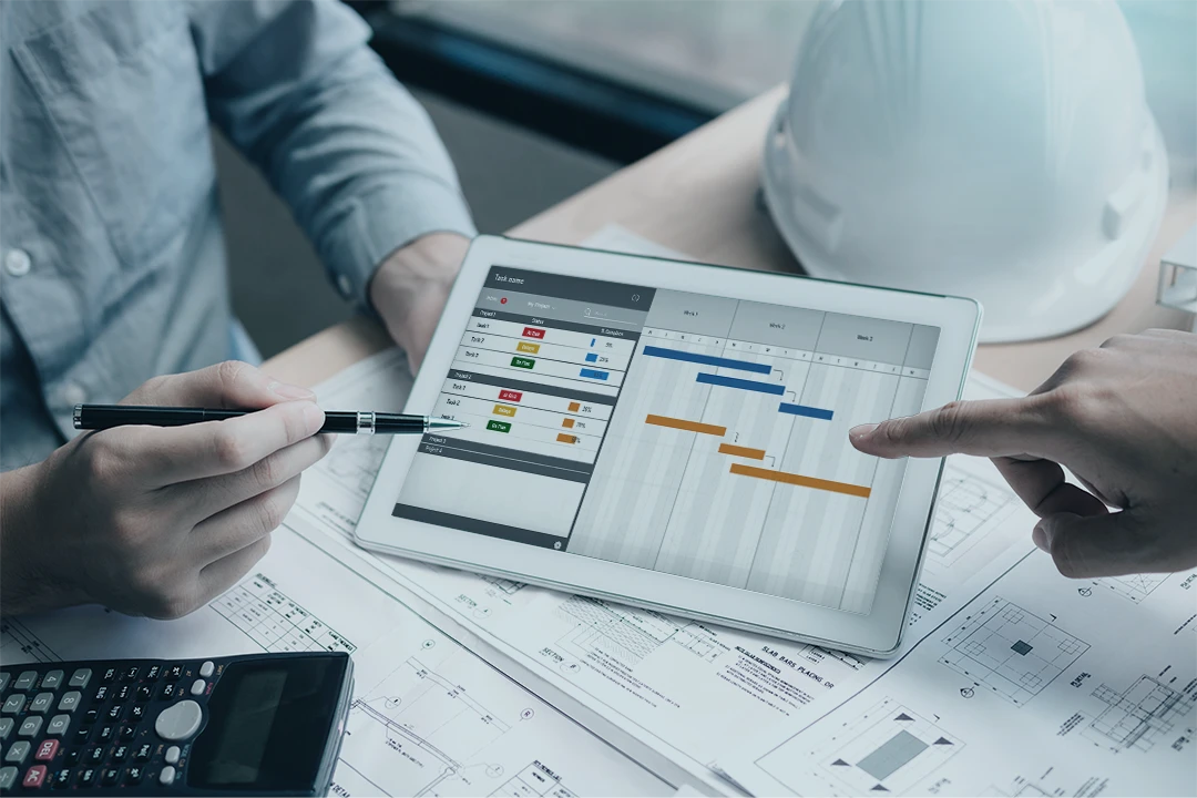 Two people review a project management Gantt chart on a tablet over construction blueprints. A calculator and a white hard hat are also on the desk.