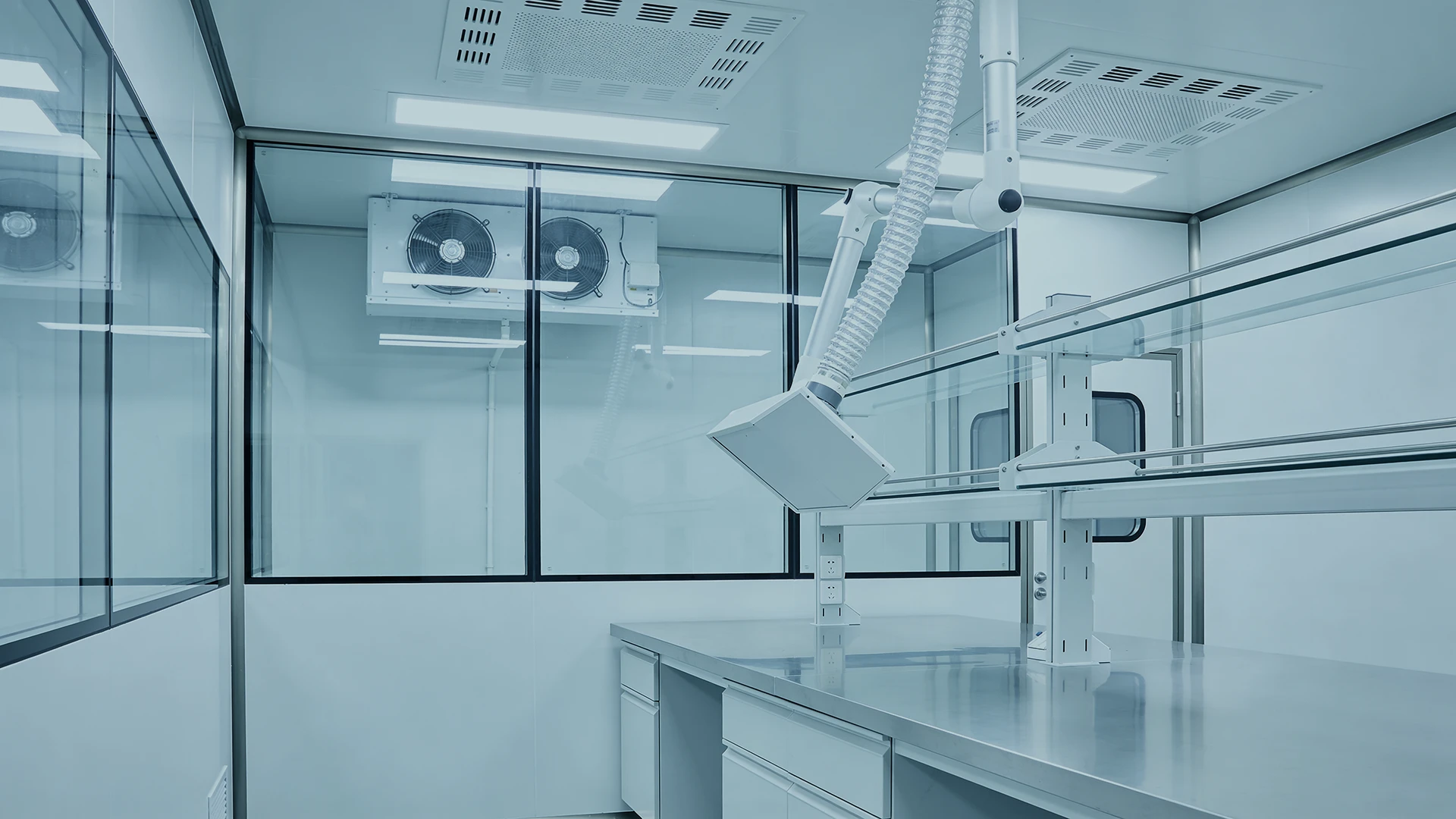 A clean, modern laboratory with stainless steel work surfaces, bright lighting, large ventilation fans, glass windows, and an adjustable exhaust arm hanging over the workstation.