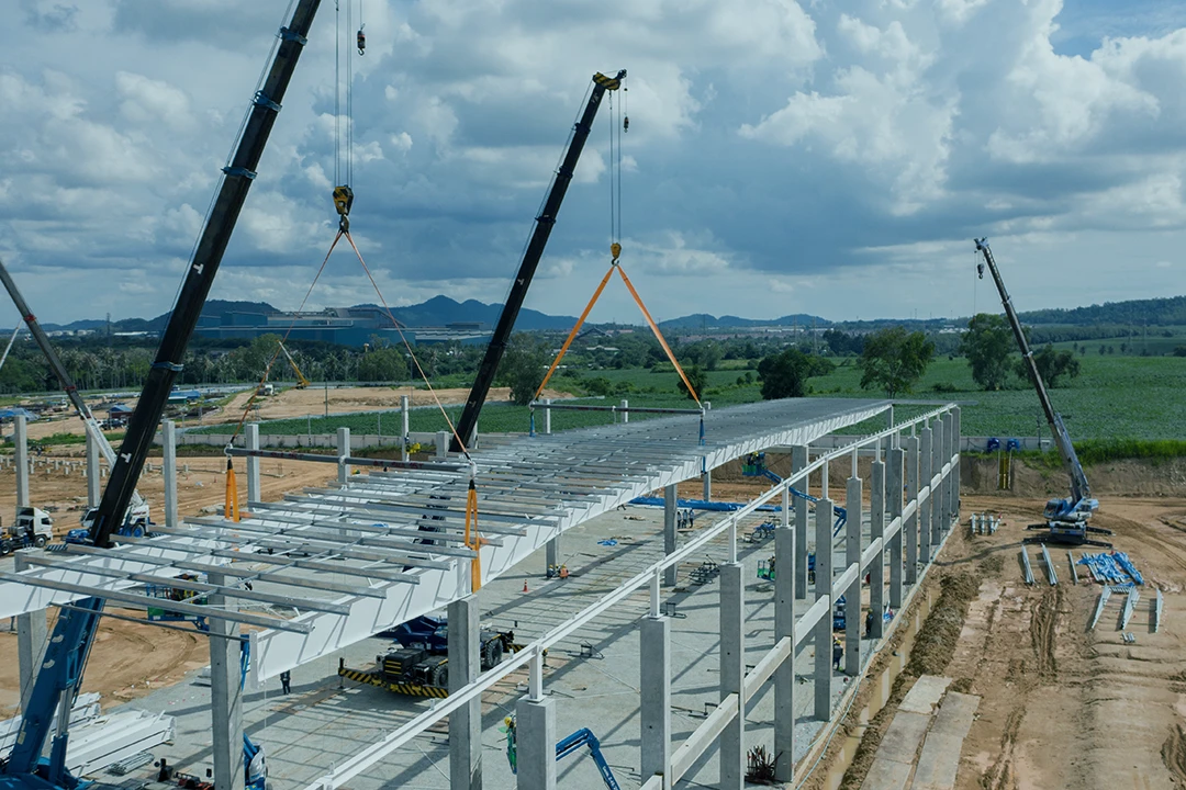 Three cranes lift steel beams to assemble a large industrial building frame at a construction site, with green fields and hills visible in the background under a partly cloudy sky.