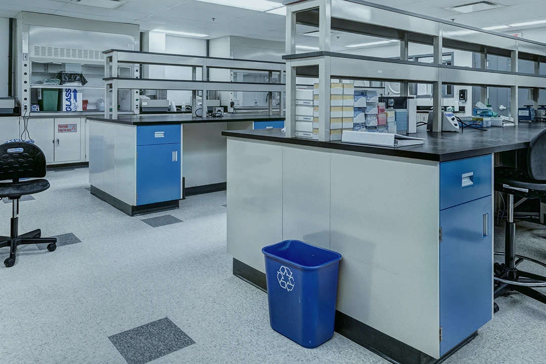 A modern laboratory with blue and white workstations, shelves, chairs, lab equipment, and a blue recycling bin on the floor. The lab is clean, well-lit, and organized, with various supplies on the counters.