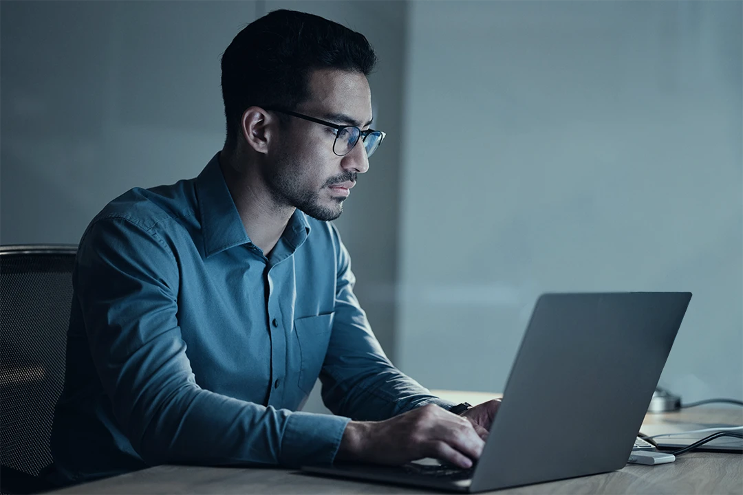 A man wearing glasses and a blue shirt sits at a desk, working intently on a laptop in a dimly lit room.