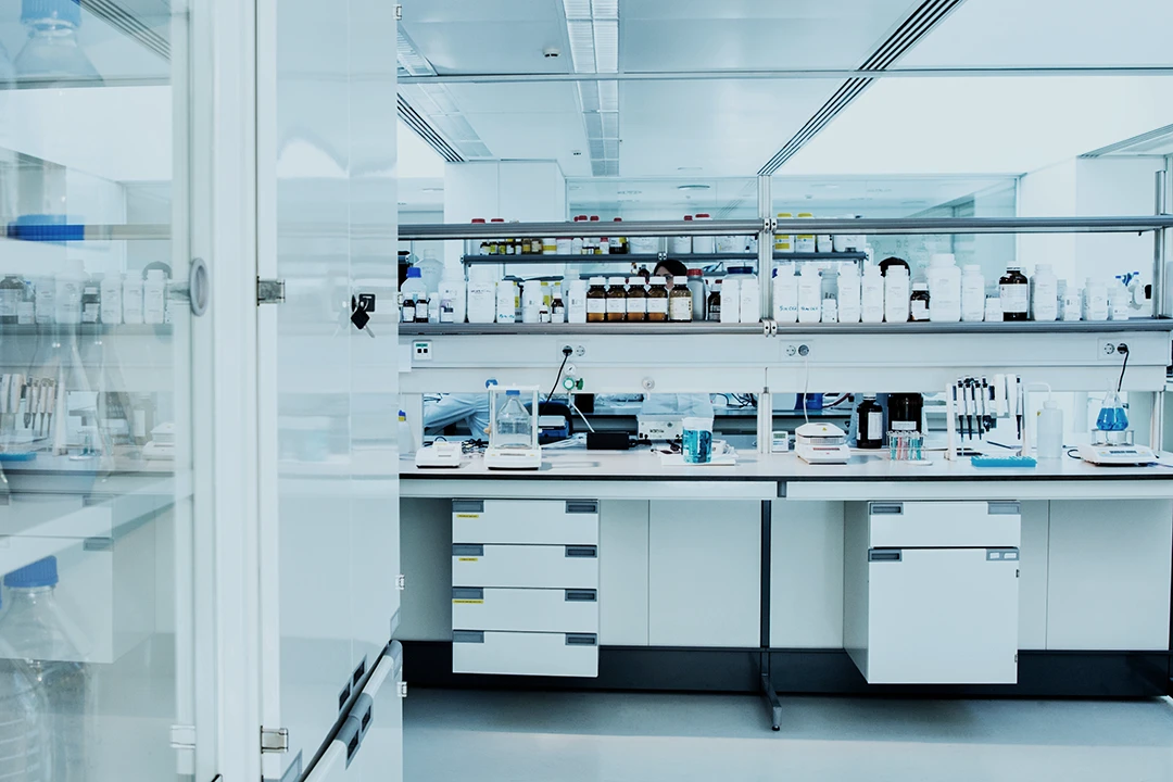 A modern, brightly lit laboratory with shelves lined with bottles and equipment, white cabinets, and workstations neatly organized for experiments.