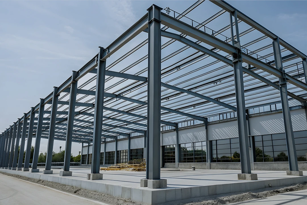 A steel frame structure of an industrial building is under construction, with beams and columns visible under a clear blue sky. Some construction materials are stacked inside the site.