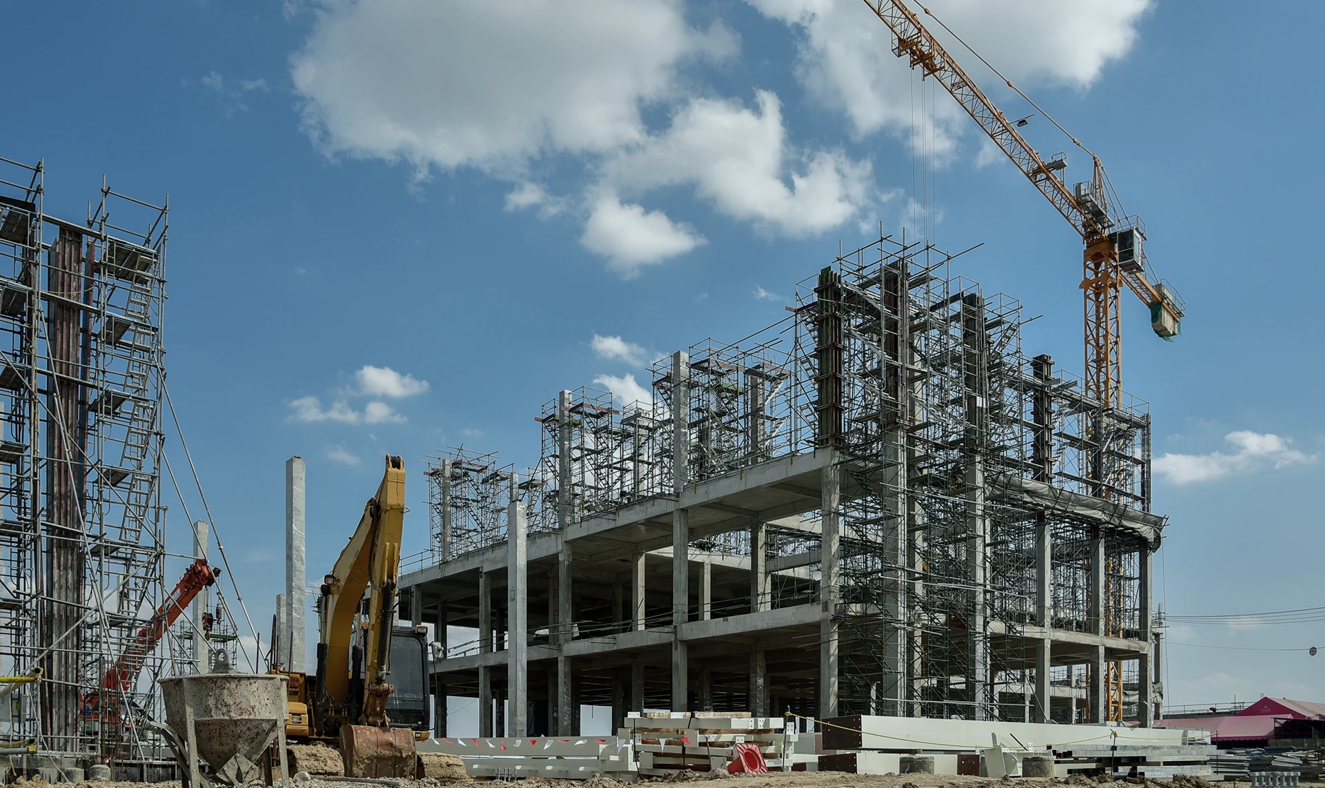 A multi-story building under construction with scaffolding, cranes, and construction vehicles against a blue sky with clouds.