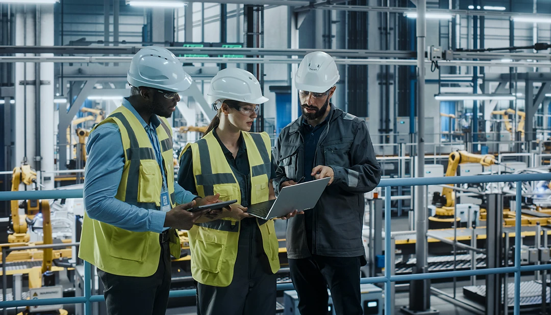 Three engineers wearing safety helmets and vests stand in a modern factory, discussing something on a laptop. Robotic machinery and industrial equipment are visible in the background.
