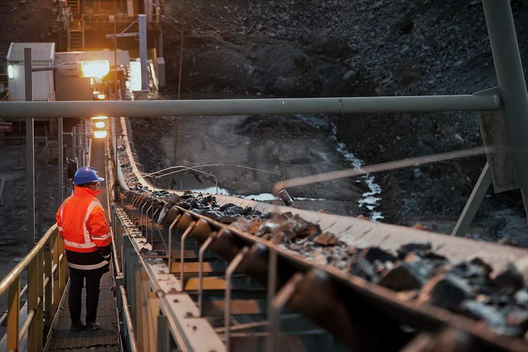 A worker in a blue helmet and orange jacket stands near a conveyor belt transporting rocks at an industrial mining site.