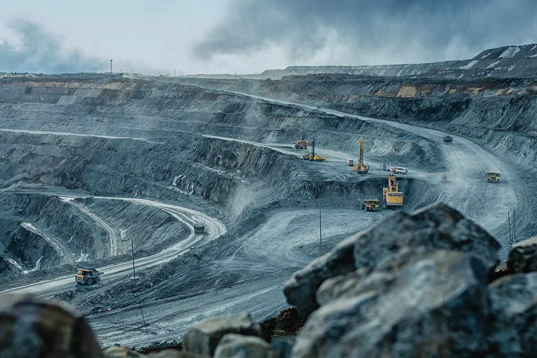 Wide view of an open-pit mine with winding roads and heavy machinery working on different levels, surrounded by grey rocky terrain under cloudy skies.