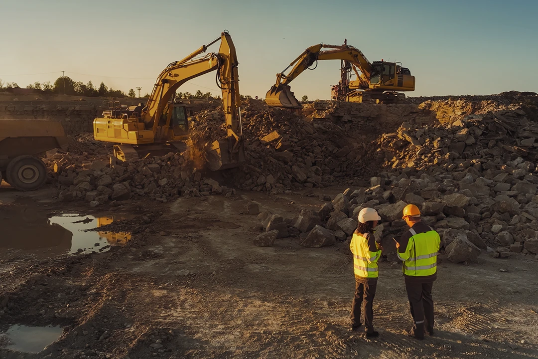 Two people in safety vests and helmets stand on rocky ground, talking, while two excavators move rocks and dirt nearby at a construction or mining site during sunset.