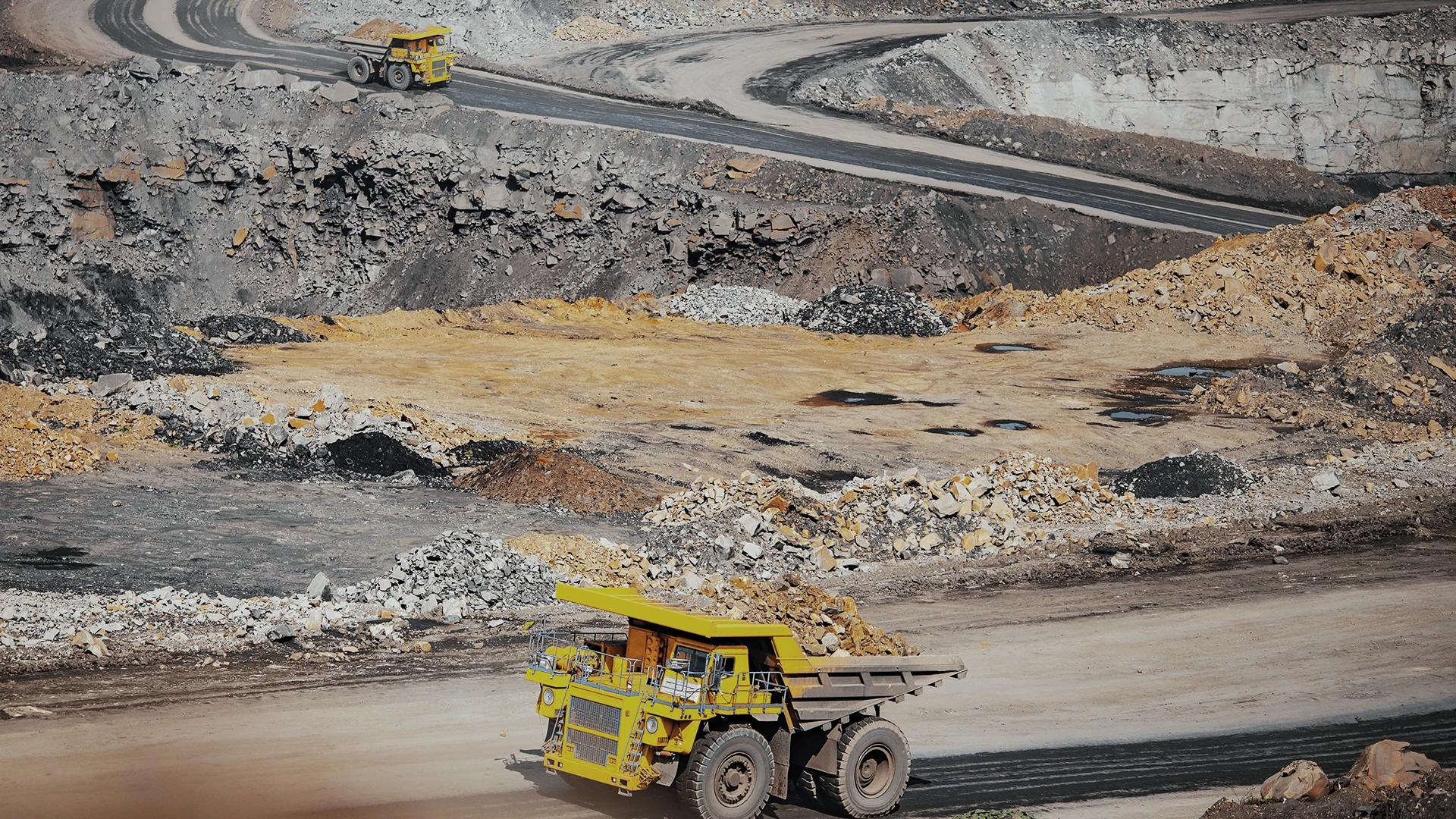 Two large yellow dump trucks operate at different levels in an open-pit mining site, surrounded by rocky terrain and dirt roads.