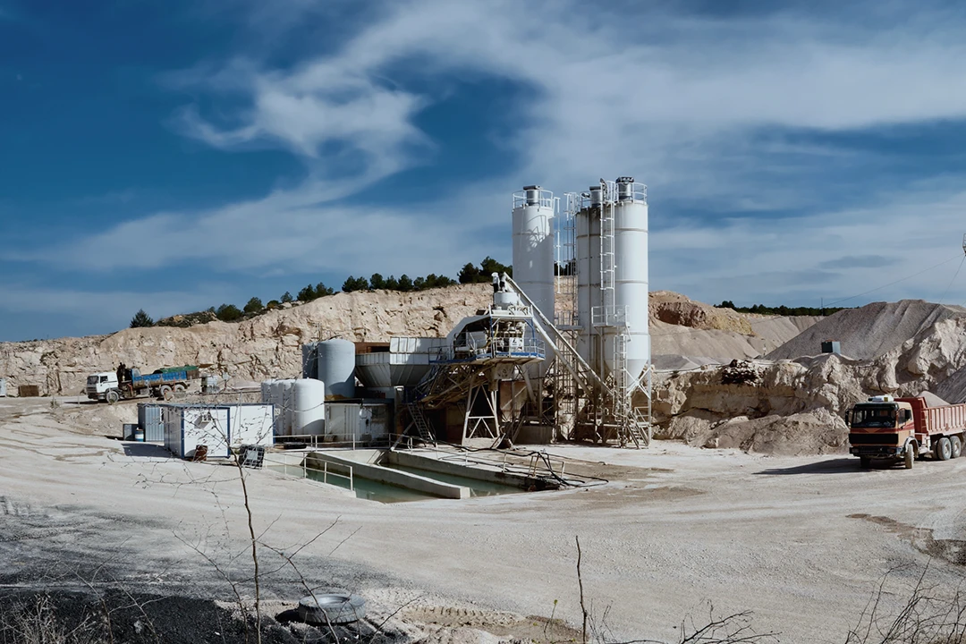 Industrial site with tall silos, machinery, trucks, and piles of sand or gravel. Blue sky with clouds in the background. Desert-like, rocky landscape surrounds the facility.