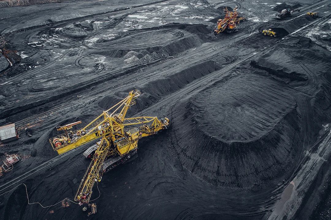Aerial view of a large open-pit coal mine with yellow excavators and machinery working surrounded by dark piles of coal.