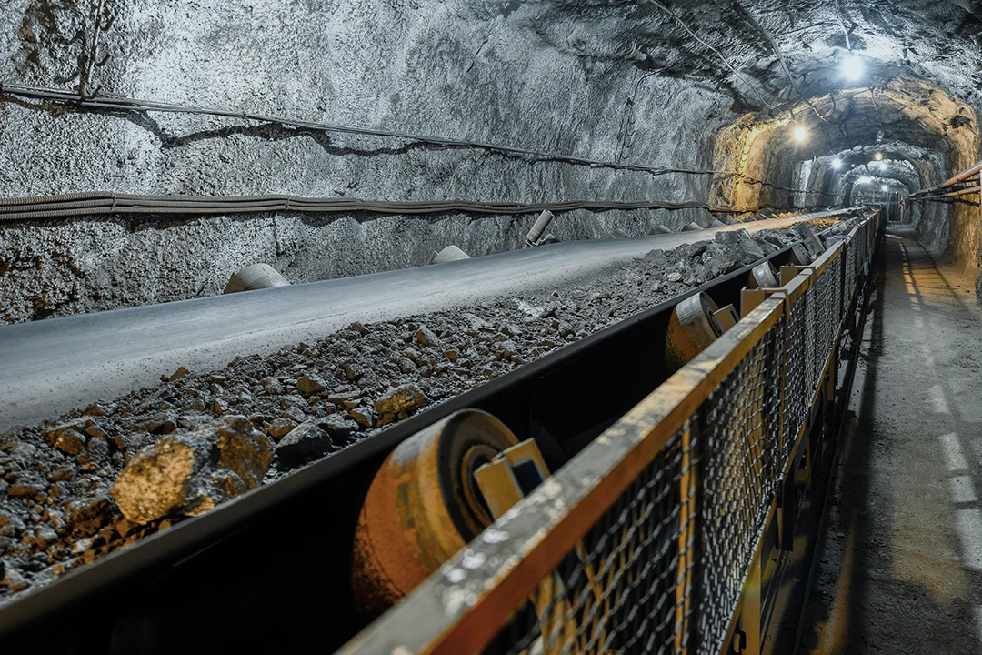 A conveyor belt transports mined rocks through an underground tunnel, with lights illuminating the rocky walls and ceiling.