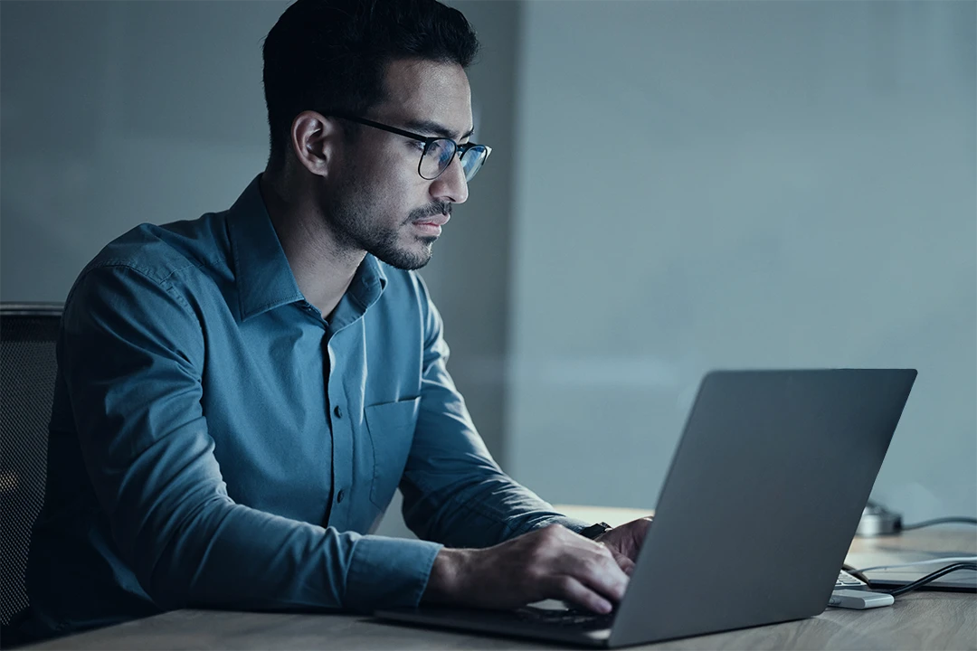 A man wearing glasses and a blue shirt sits at a desk, typing on a laptop.