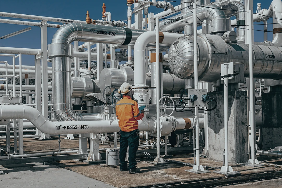 A worker in a hard hat and orange jacket inspects pipes and valves at an industrial facility with large metal pipelines and equipment under a clear blue sky.