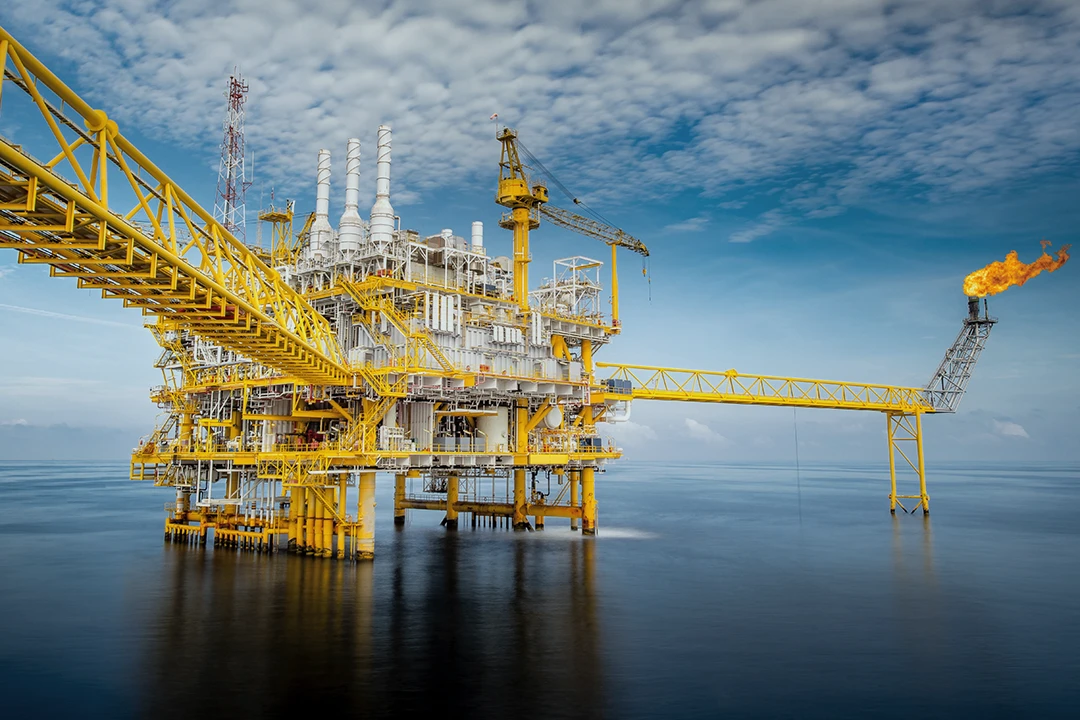 A large yellow offshore oil rig stands in calm ocean water under a partly cloudy sky, with a gas flare burning at the end of a platform extending over the sea.