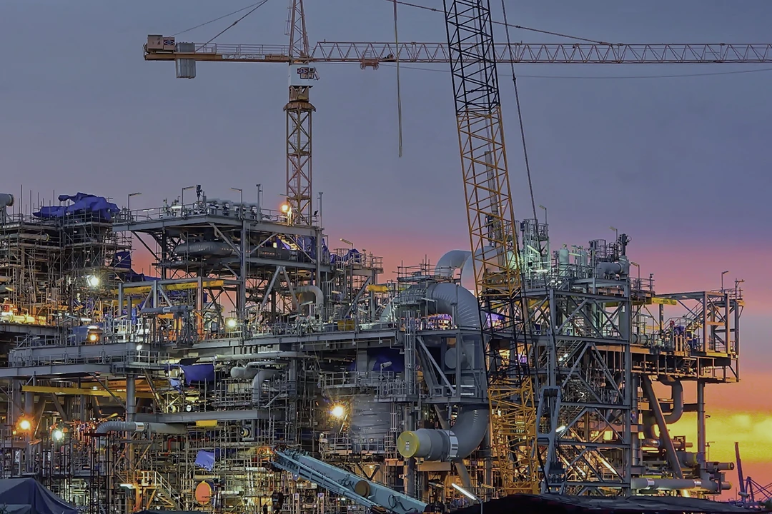 Large industrial construction site with cranes and scaffolding, illuminated by lights, set against a colorful sunset sky in the background. Various pipes and structures are visible throughout the complex scene.