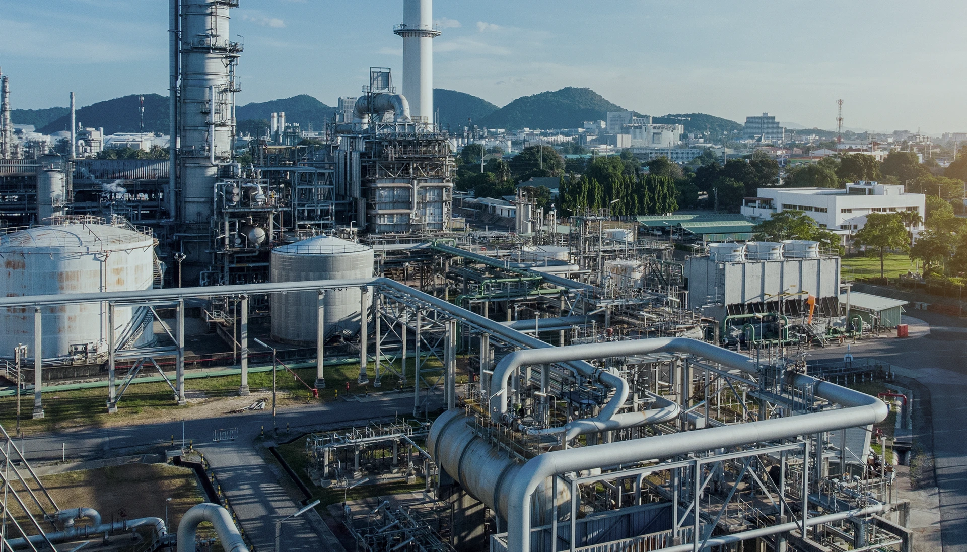 Industrial refinery complex with large storage tanks, metal pipes, and distillation towers. Buildings and green hills are visible in the background under a clear sky.
