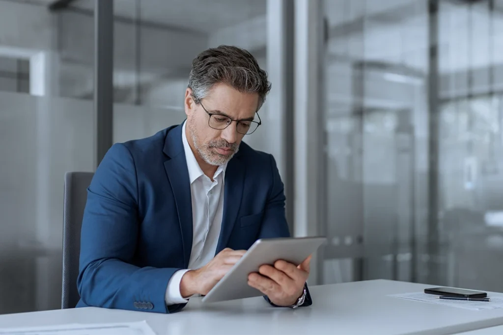 A man in a blue suit and glasses sits at a desk in an office, using a tablet. Papers and a smartphone are on the desk. The background shows glass walls.
