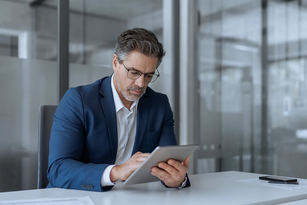 A man in a blue suit and glasses sits at a desk in an office, using a tablet. Papers and a smartphone are on the desk. The background shows glass walls.