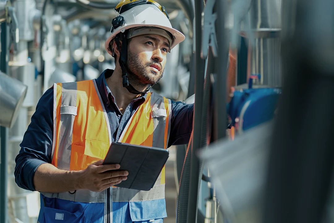 A construction worker wearing a hard hat and orange safety vest inspects equipment while holding a tablet, surrounded by industrial machinery.