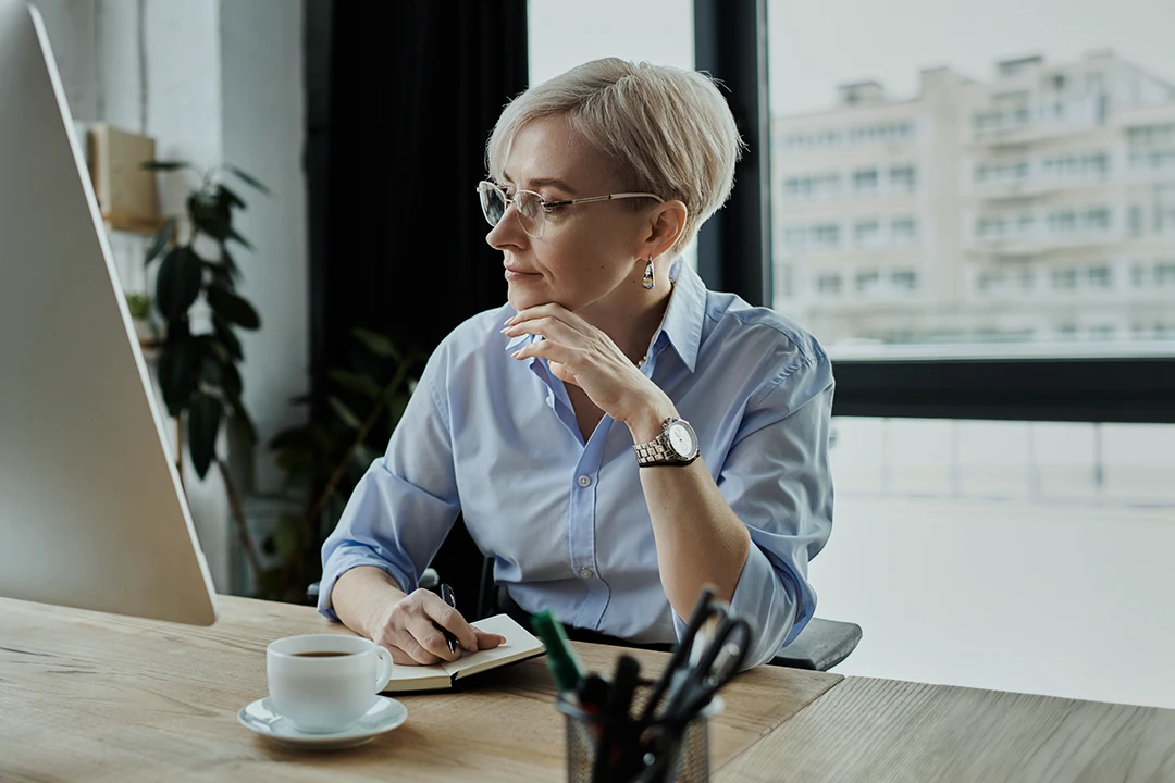 A person with short blonde hair and glasses sits at a desk by a window, looking at a computer monitor while holding a pen and notebook. There’s a cup of coffee and office supplies on the wooden desk.