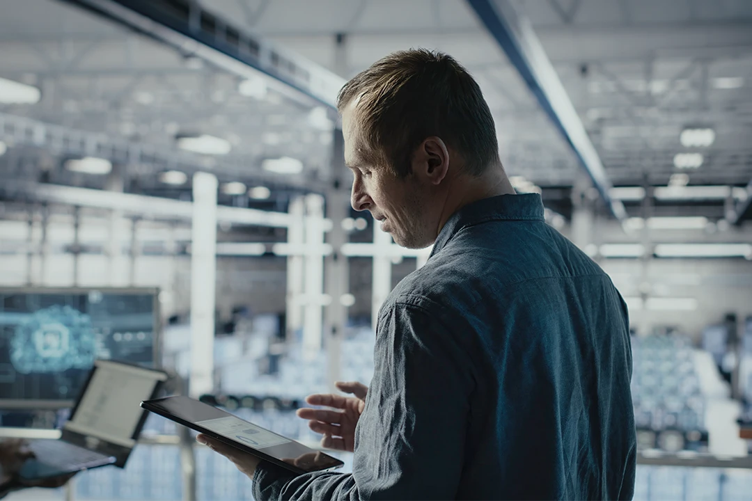 A man in a denim shirt uses a tablet in a modern, well-lit industrial or tech workspace with digital screens and equipment in the background.
