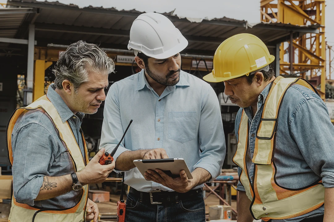 Three construction workers wearing safety vests and helmets stand together at a job site. One holds a radio, another uses a tablet, and the third listens attentively. Equipment and materials are visible in the background.