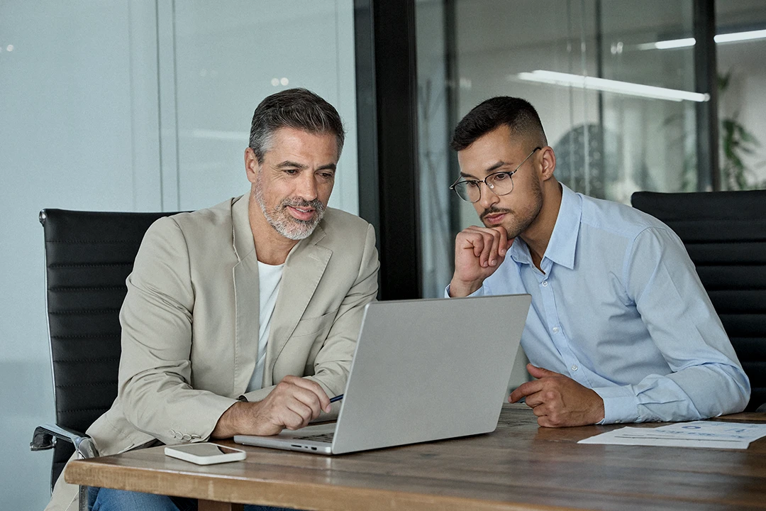 Two men in business attire sit at a desk, looking at a laptop screen and discussing something. Papers and a smartphone are on the wooden table. The setting appears to be a modern office.