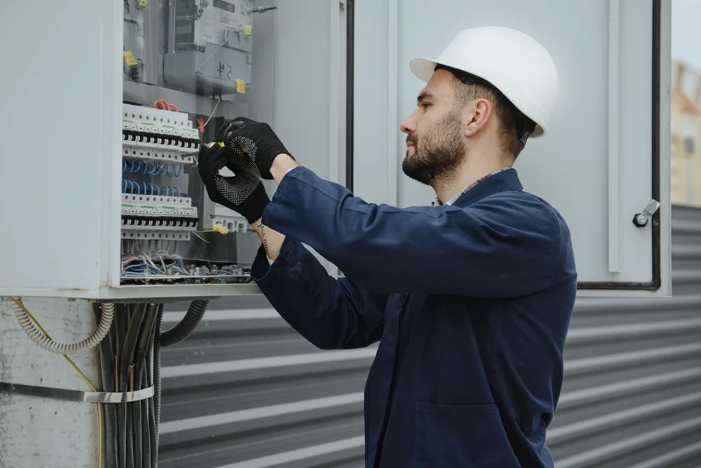 A man wearing a white hard hat, black gloves, and a dark jacket works on an open electrical panel mounted on a wall, handling wires and switches.