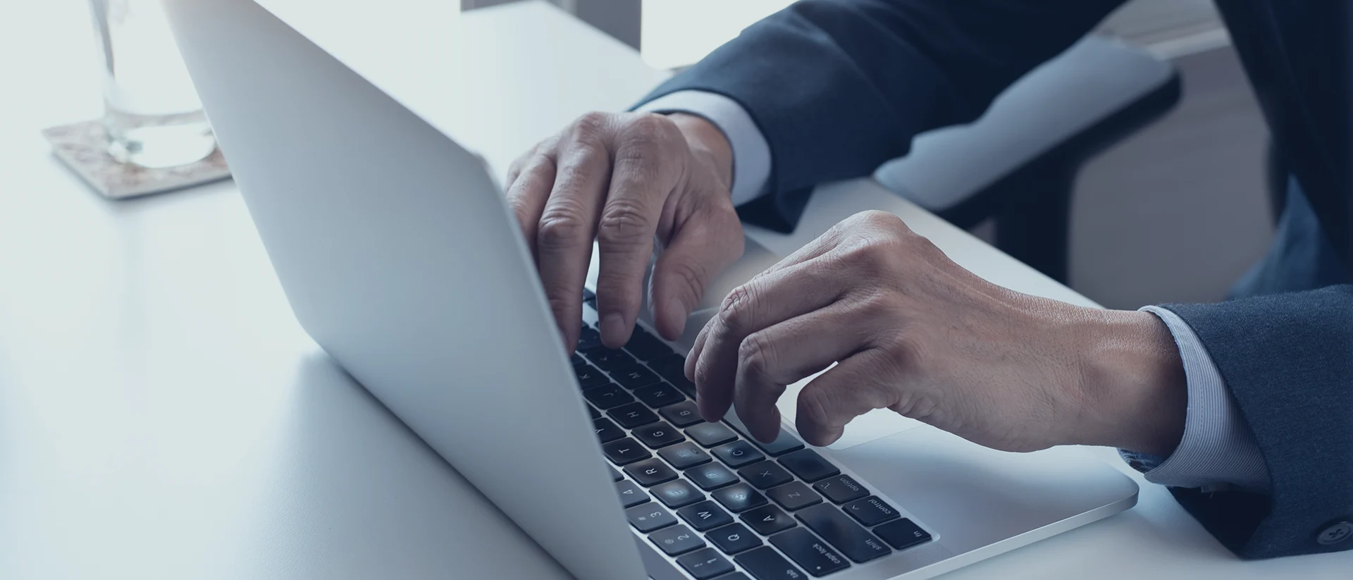 A person in a suit types on a silver laptop at a white desk, with a glass of water in the background.