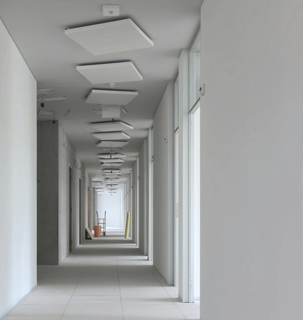 A long, empty corridor in a building under construction, featuring tiled floors, white walls, and a row of square light fixtures mounted on the ceiling.
