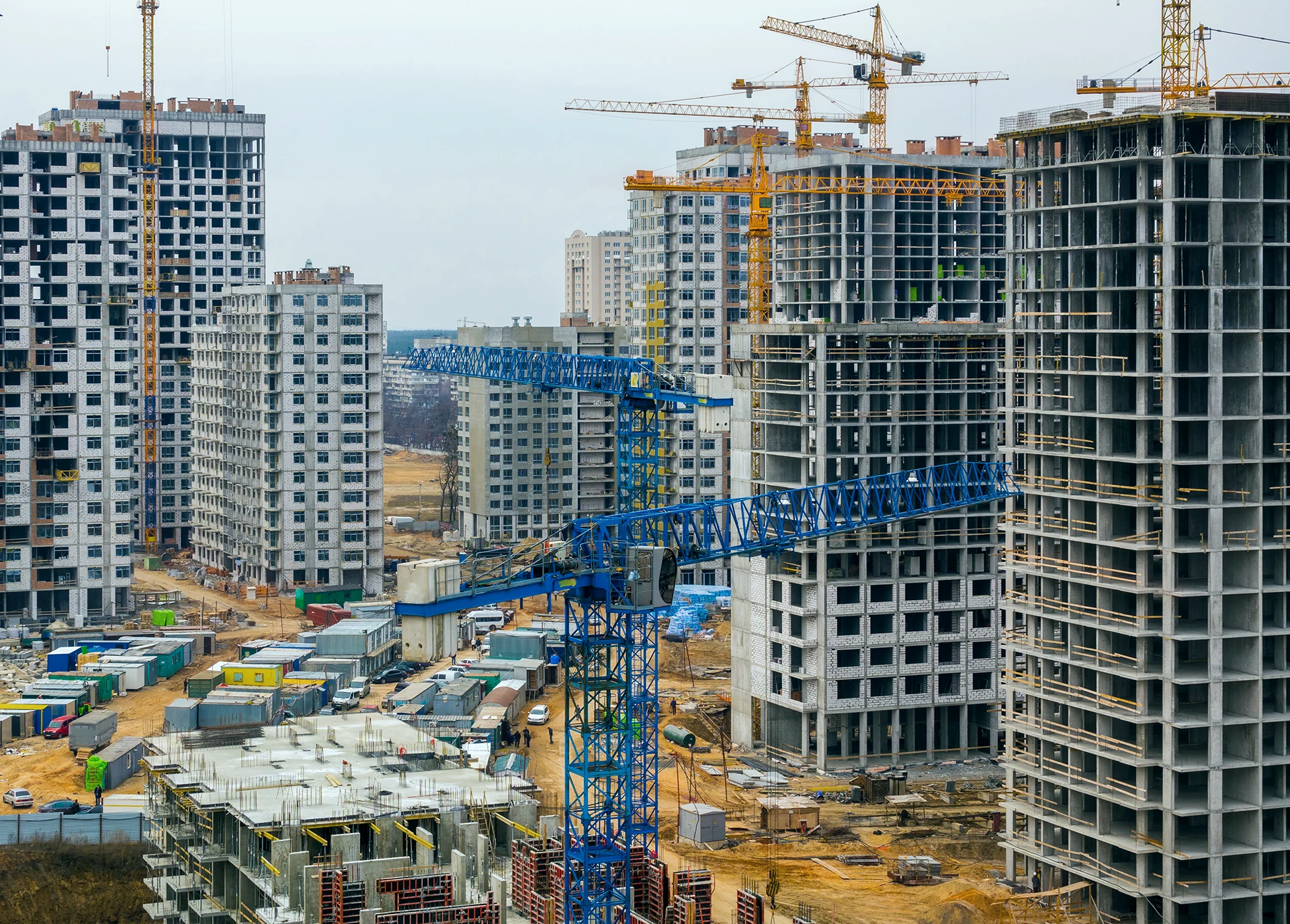Several high-rise buildings under construction with cranes and construction equipment on a large site, surrounded by unfinished structures and materials under a cloudy sky.