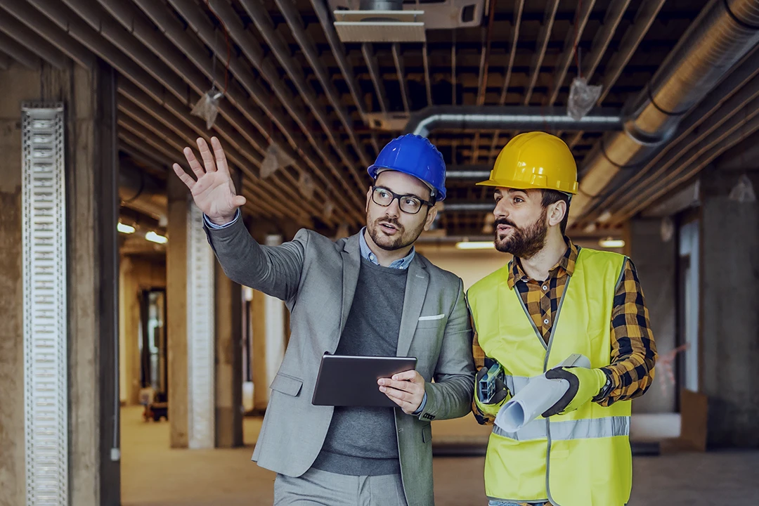 Two men in hard hats, one in a suit with a tablet and the other in a safety vest holding blueprints, stand in a building under construction, discussing plans while one gestures forward.
