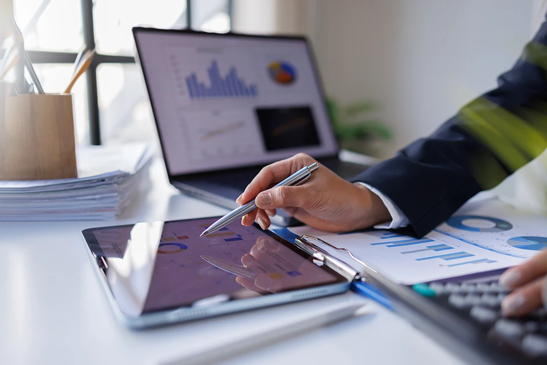 Person analyzing financial data on a tablet, with charts and graphs visible on both the tablet and a laptop screen. Papers and a calculator are on the desk, suggesting work on business or finance analysis.