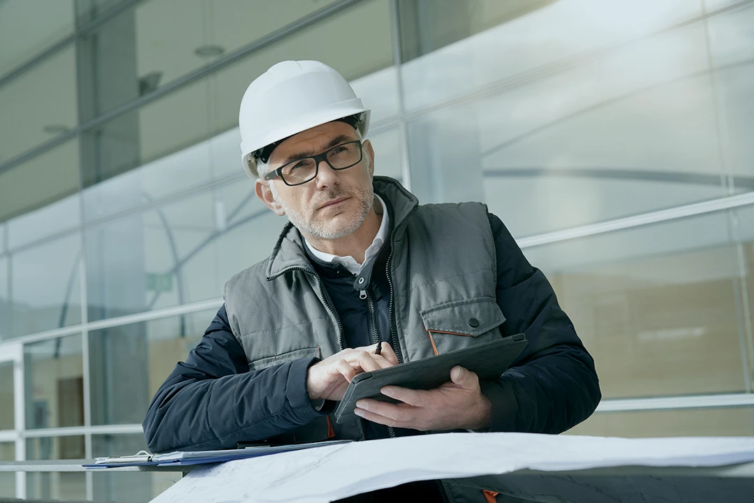 A man wearing a white hard hat and glasses looks at a tablet while sitting at a table with blueprints, inside a modern building with large windows.