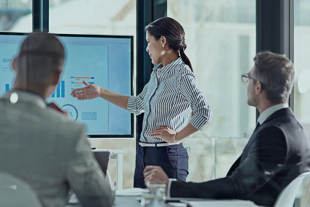 A woman stands and points at a screen displaying charts during a business meeting, while two men in suits sit at a conference table and listen attentively.