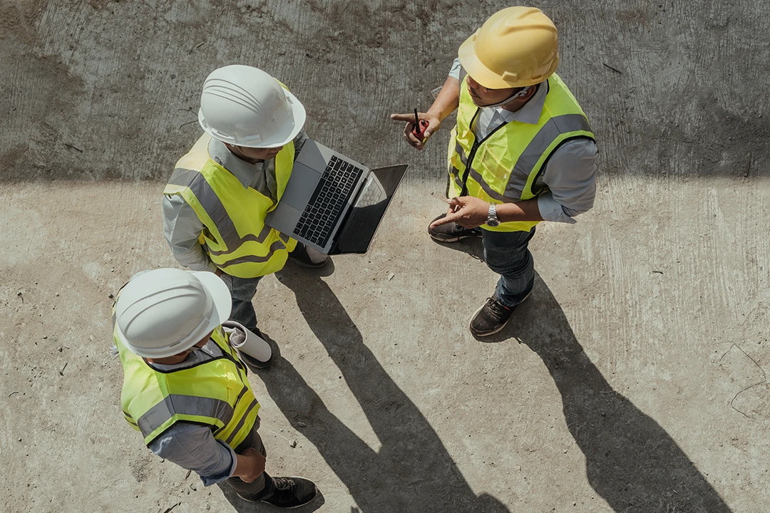 Three construction workers wearing safety vests and helmets stand together outdoors. One holds a laptop, while another gestures with a pen as they discuss something, viewed from above on a concrete surface.