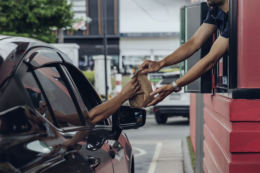 A person in a car reaches out to receive a brown paper bag from an employee at a drive-thru window.