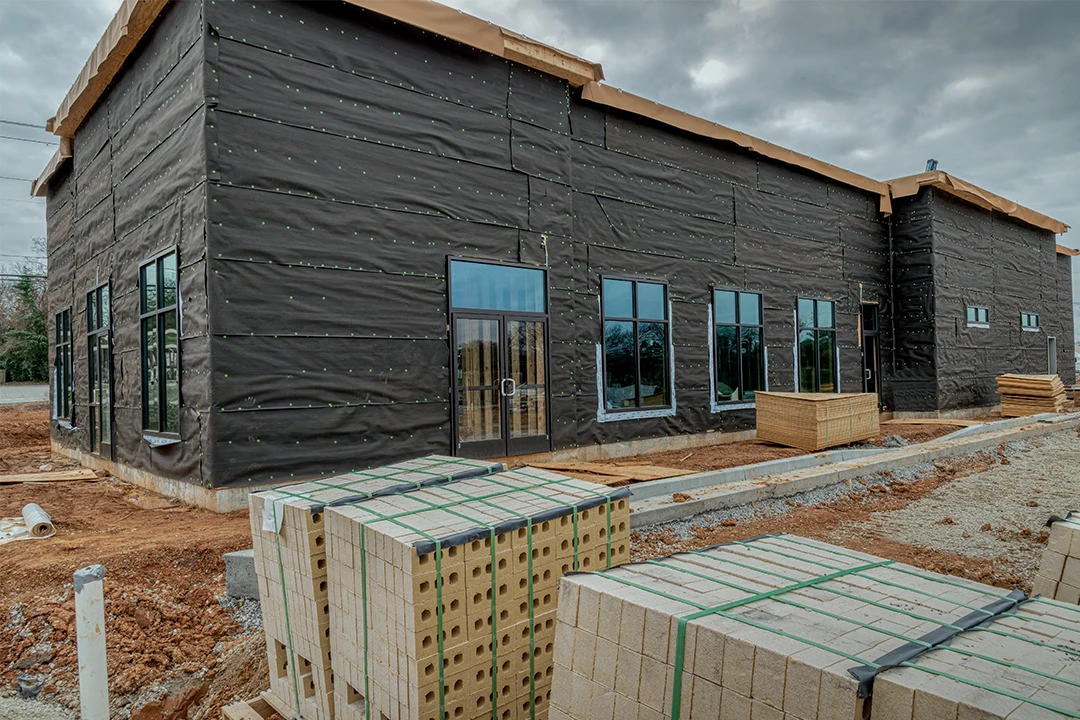 A commercial building under construction, covered in black weatherproof sheathing, with blue-framed windows and construction materials like bricks stacked in the foreground on a cloudy day.
