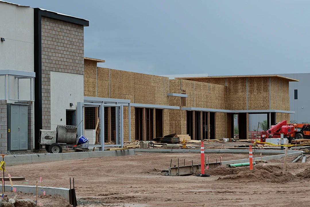 A commercial building under construction, with exposed wooden framing, construction materials, machinery, and dirt in the foreground under a cloudy sky.