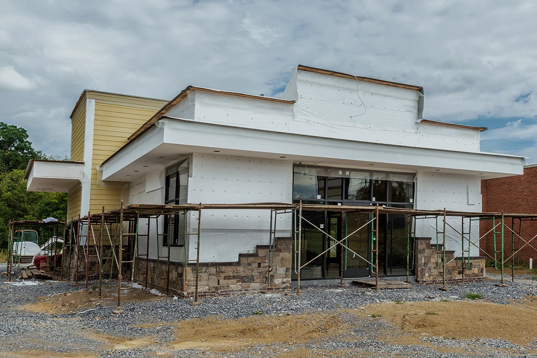 A small modern building under construction, with scaffolding around the exterior, unfinished walls, and a partially gravel-covered ground in front.